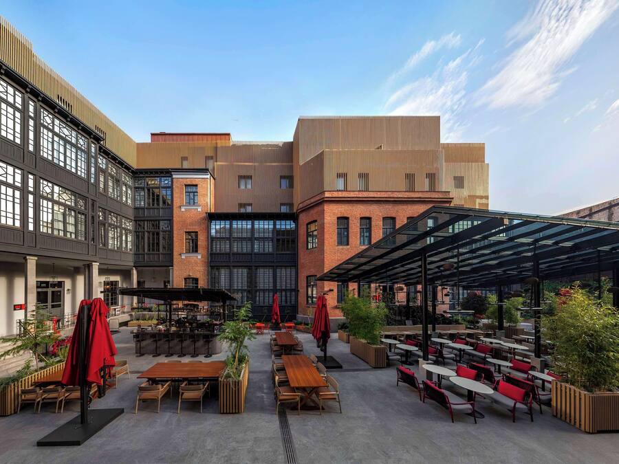 An outdoor dining area with tables and chairs, surrounded by buildings with large windows and a covered seating area.