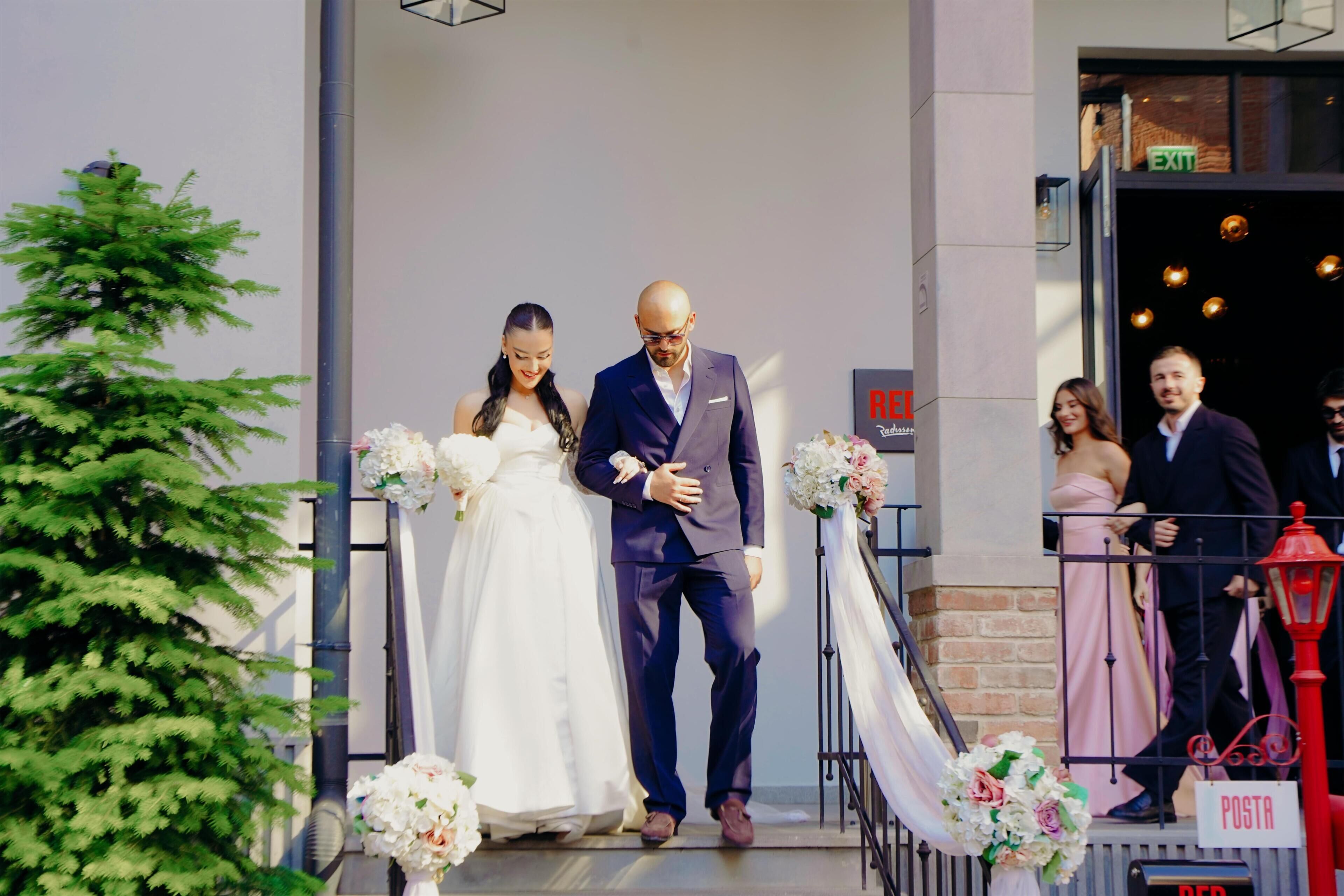 A couple exiting a building with a wedding dress and a suit, a sign reading 'POSTA,' and a red lamp post.