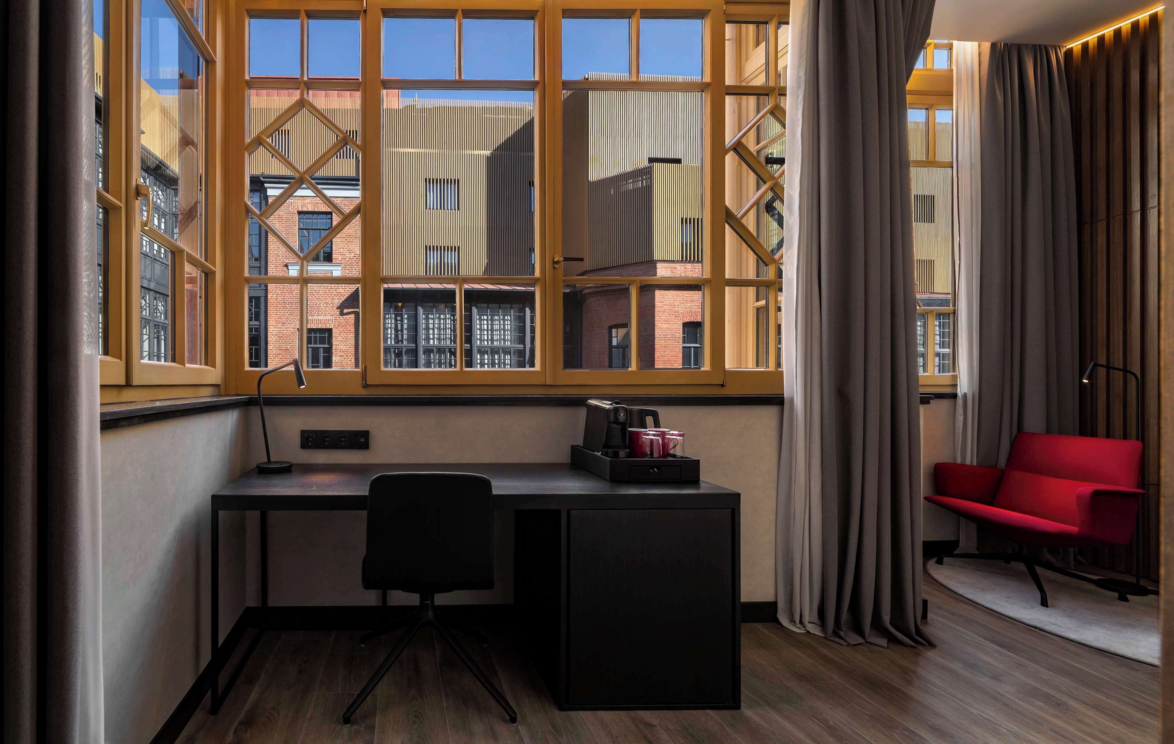 A modern room with a desk, chair, and a red sofa. A large window with curtains is visible, offering a view of buildings outside.