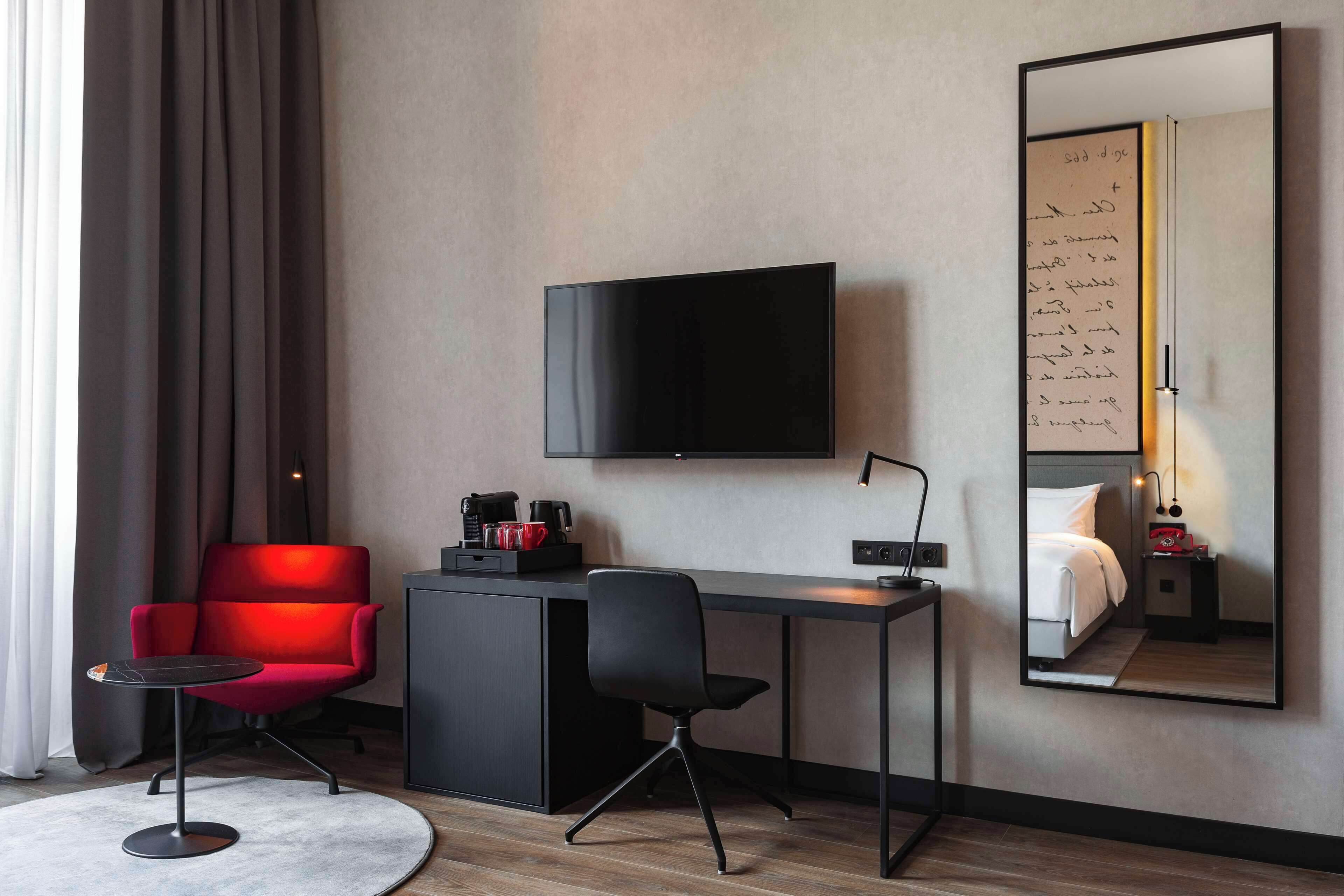 A modern hotel room with a flat-screen TV, a desk, a red chair, a black chair, and a large mirror reflecting the room.