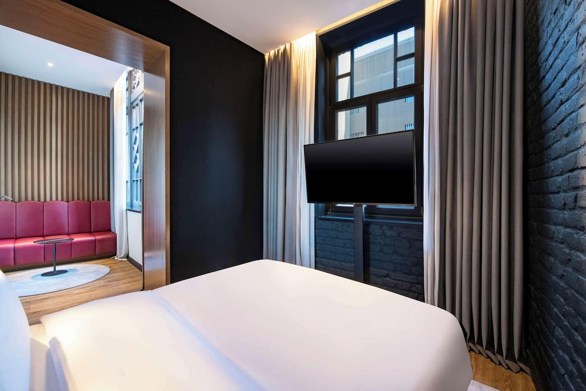 A modern hotel room with a large bed, a flat-screen TV, a red sofa, and a large window with curtains.