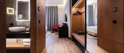 A modern hotel room with a large mirror, a wooden vanity, a bed, and a seating area with a red chair.