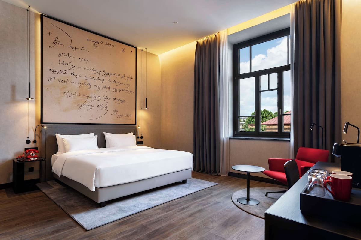 A hotel room with a large bed, a desk with a chair, a small table, and a view of the outdoors through a window.