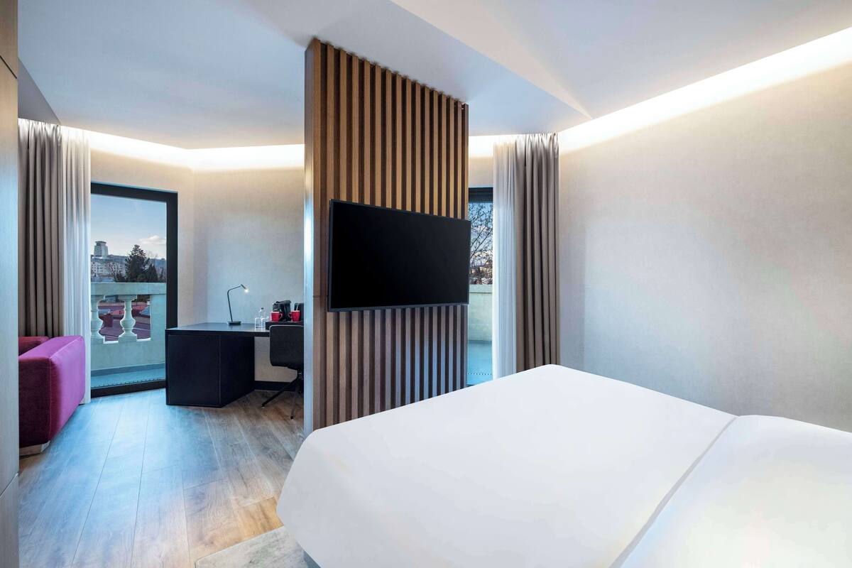 A modern hotel room with a large bed, a desk with a chair, a flat-screen TV, and a view of the city through a large window.