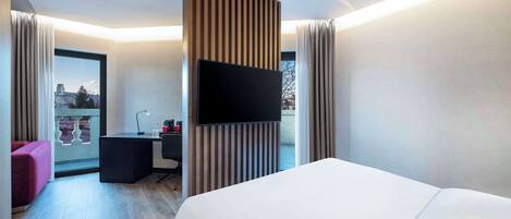 A modern hotel room with a large bed, a desk with a chair, a flat-screen TV, and a view of the city through a large window.