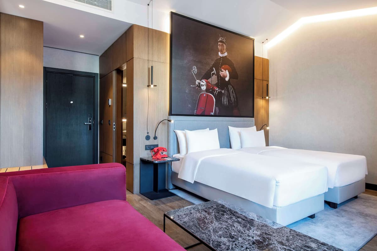 A modern hotel room with a large bed, a red sofa, a small table with a red telephone, and a wall-mounted painting.