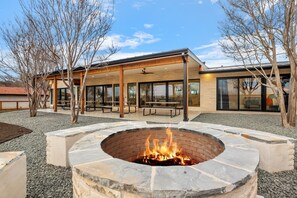 An outdoor patio area with a fire pit, picnic tables, and a covered seating area with large windows. - Guadalupe Bluff Modern House 5 Bedroom Home by RedAwning (Kerrville)