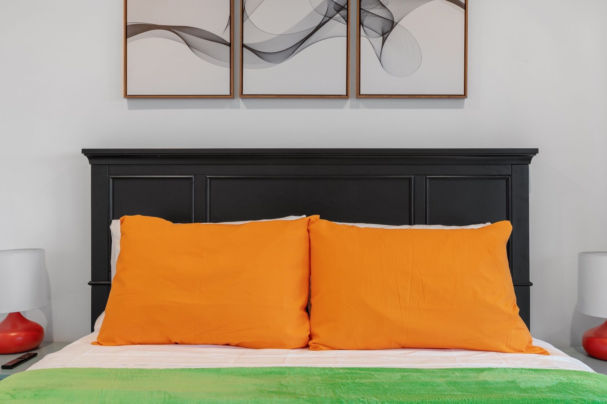A bed with two orange pillows, a black headboard, and a green blanket. There are two red lamps on either side of the bed.