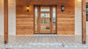 Une porte en bois avec des panneaux de verre, un hall d’entrée carrelé et deux lampes fixées au mur.