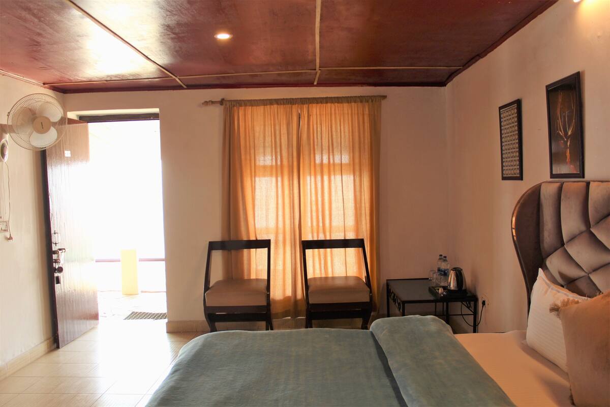 Habitación de hotel con cama, dos sillas, una mesita con hervidor, un ventilador y una ventana con cortinas.