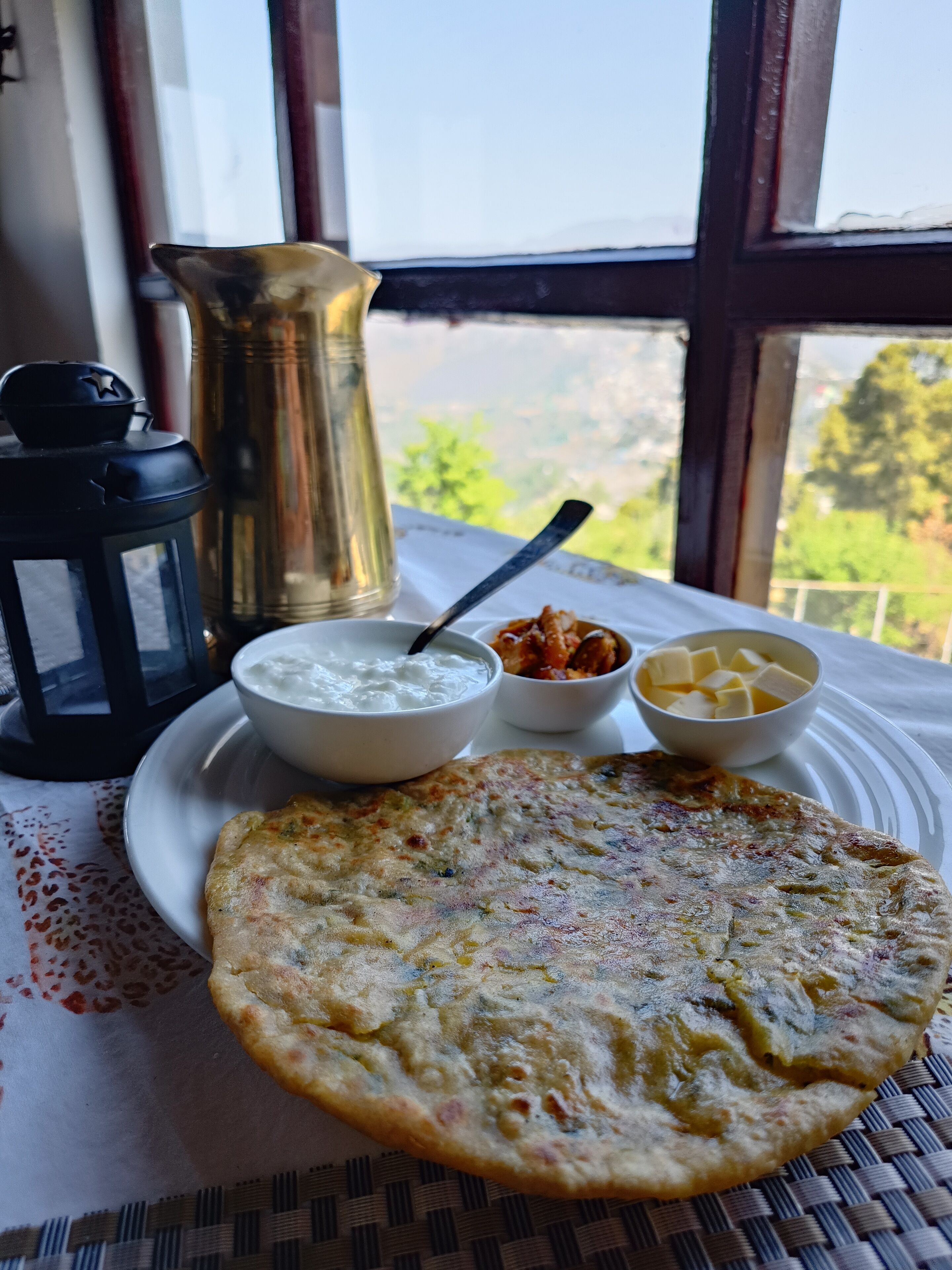 A table with a flatbread, a bowl of yogurt, and a bowl of butter, set on a tablecloth near a window with a view of greenery.