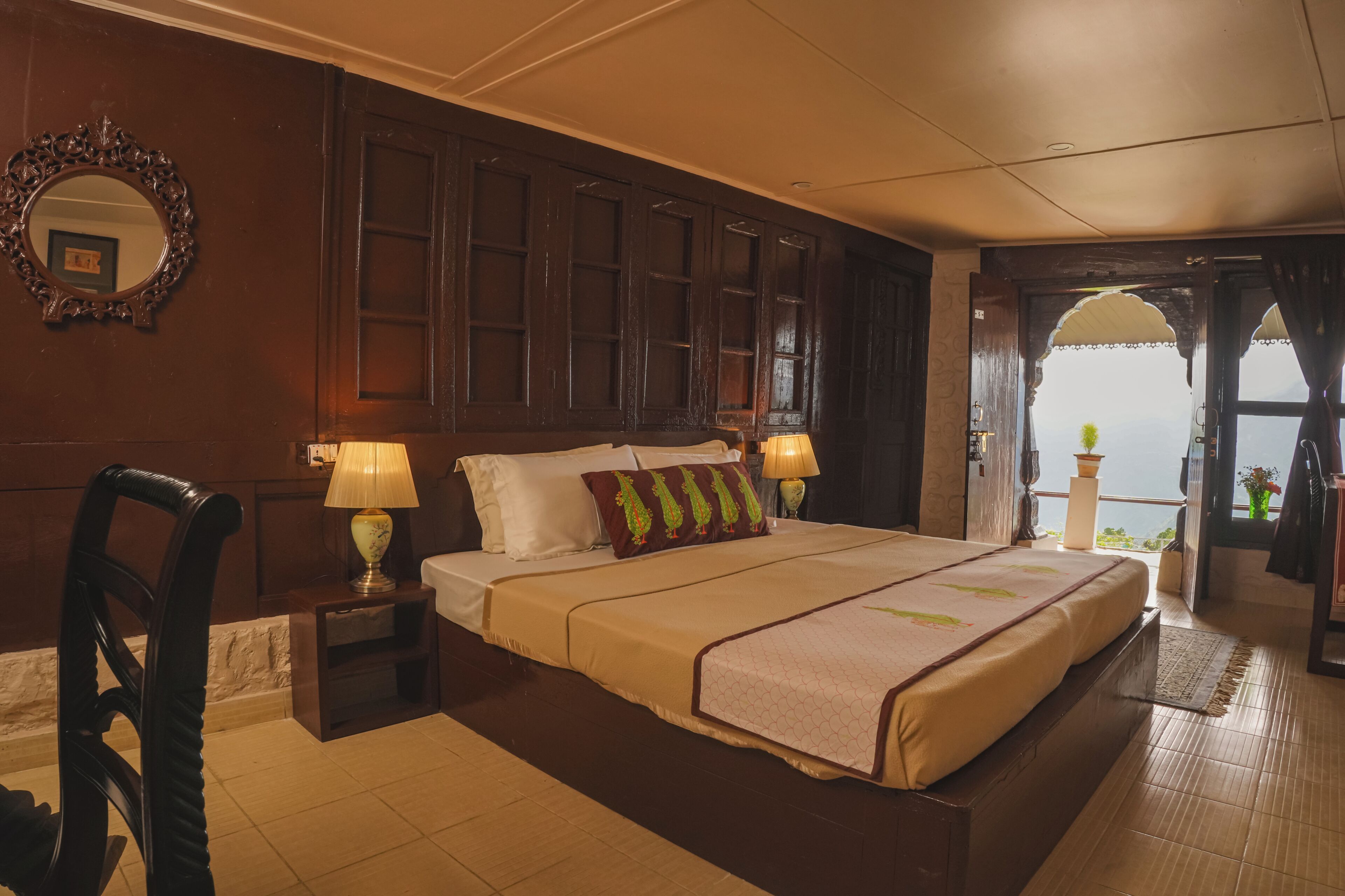 Quarto de hotel com uma cama grande, cabeceira de madeira, mesa de cabeceira com abajur, uma cadeira e vista para o mar através de uma janela em arco.
