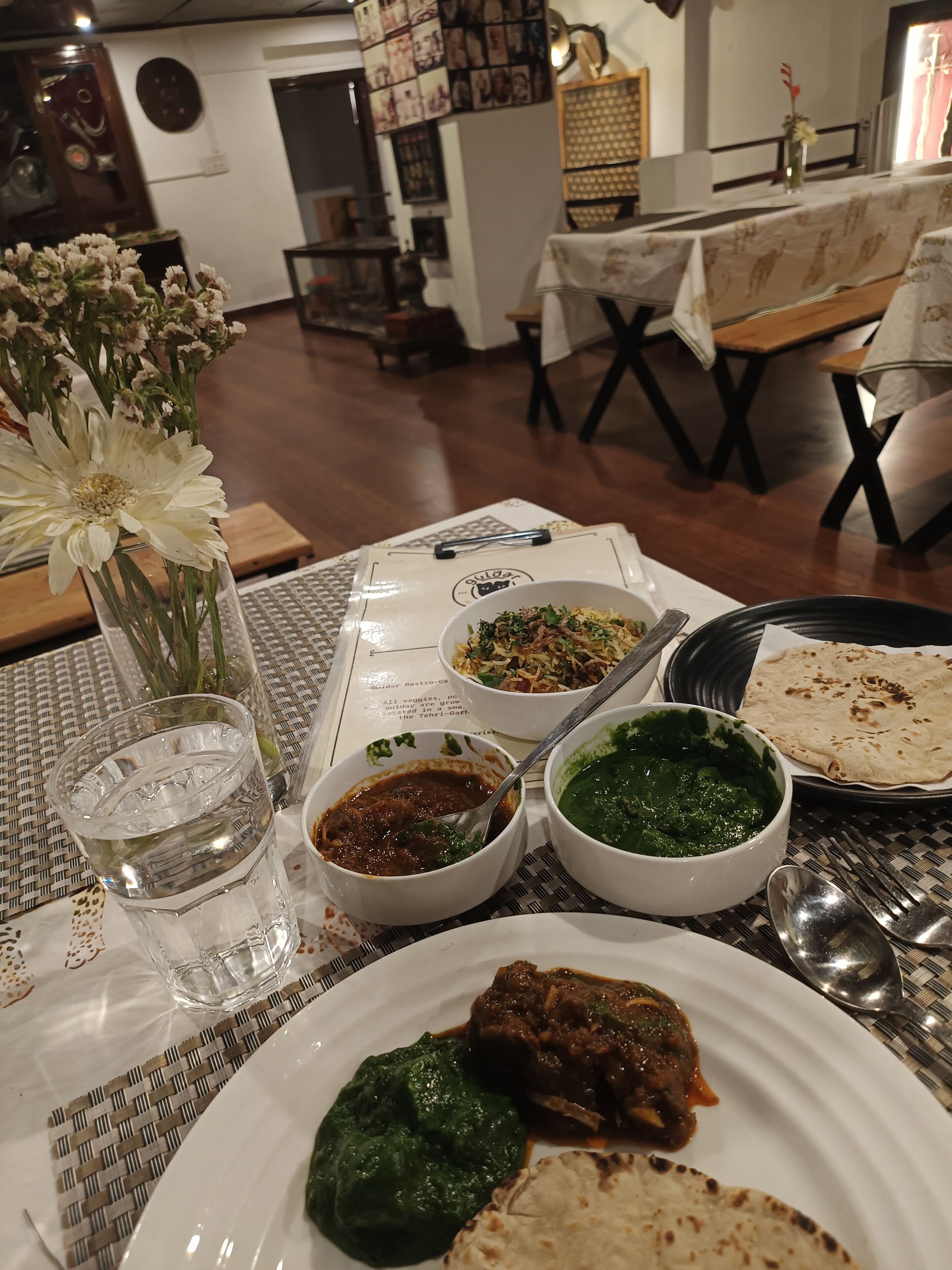 Una mesa de comedor con varios platos, incluyendo un plato con pan plano, un tazón de curry verde y un tazón de curry rojo. También hay un vaso de agua y un florero con flores.