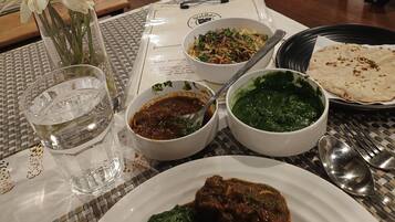 Una mesa de comedor con varios platos, incluyendo un plato con pan plano, un tazón de curry verde y un tazón de curry rojo. También hay un vaso de agua y un florero con flores.