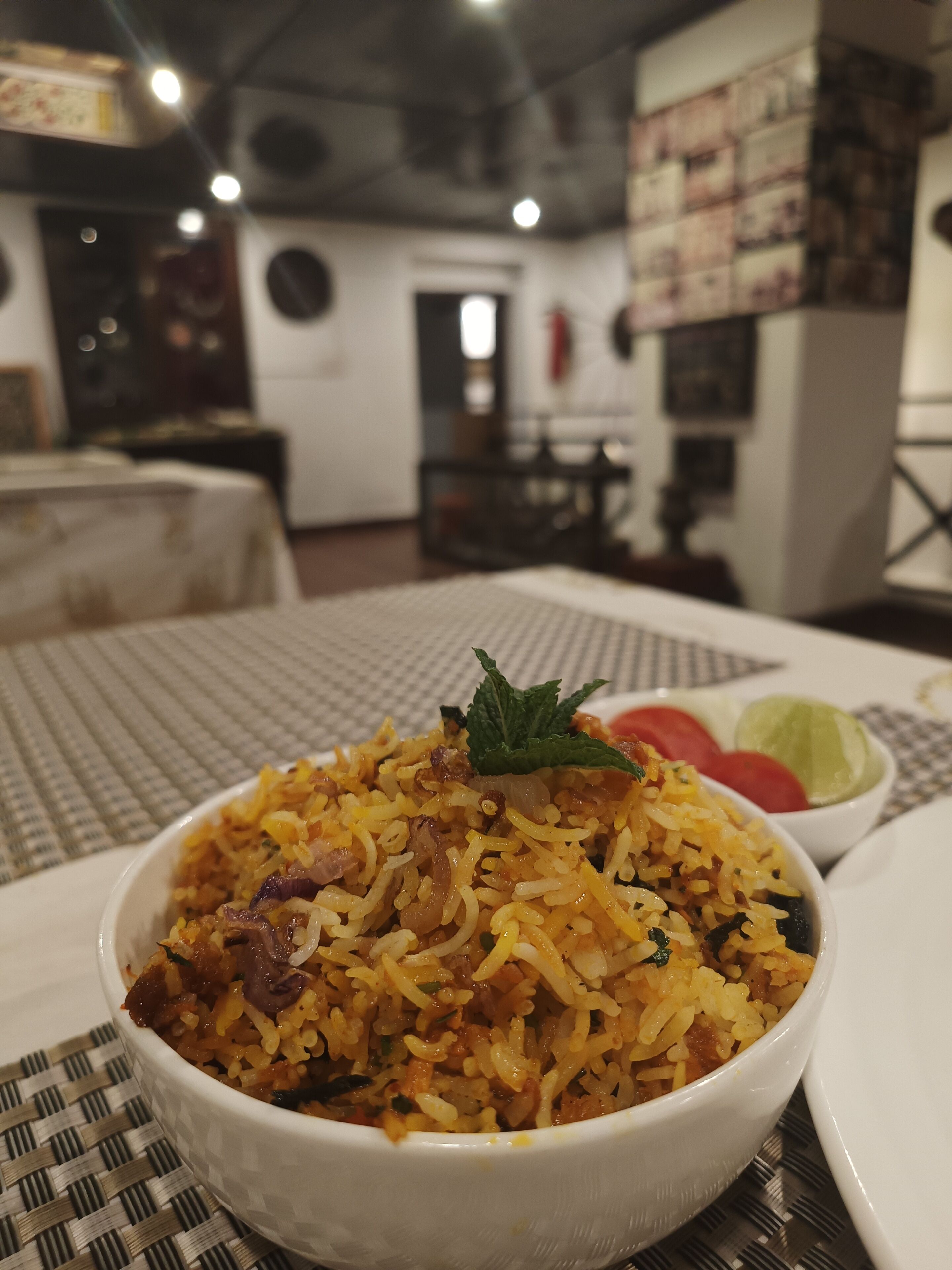 A bowl of biryani with a mint leaf garnish, a side of sliced tomatoes, and a small bowl of raita.