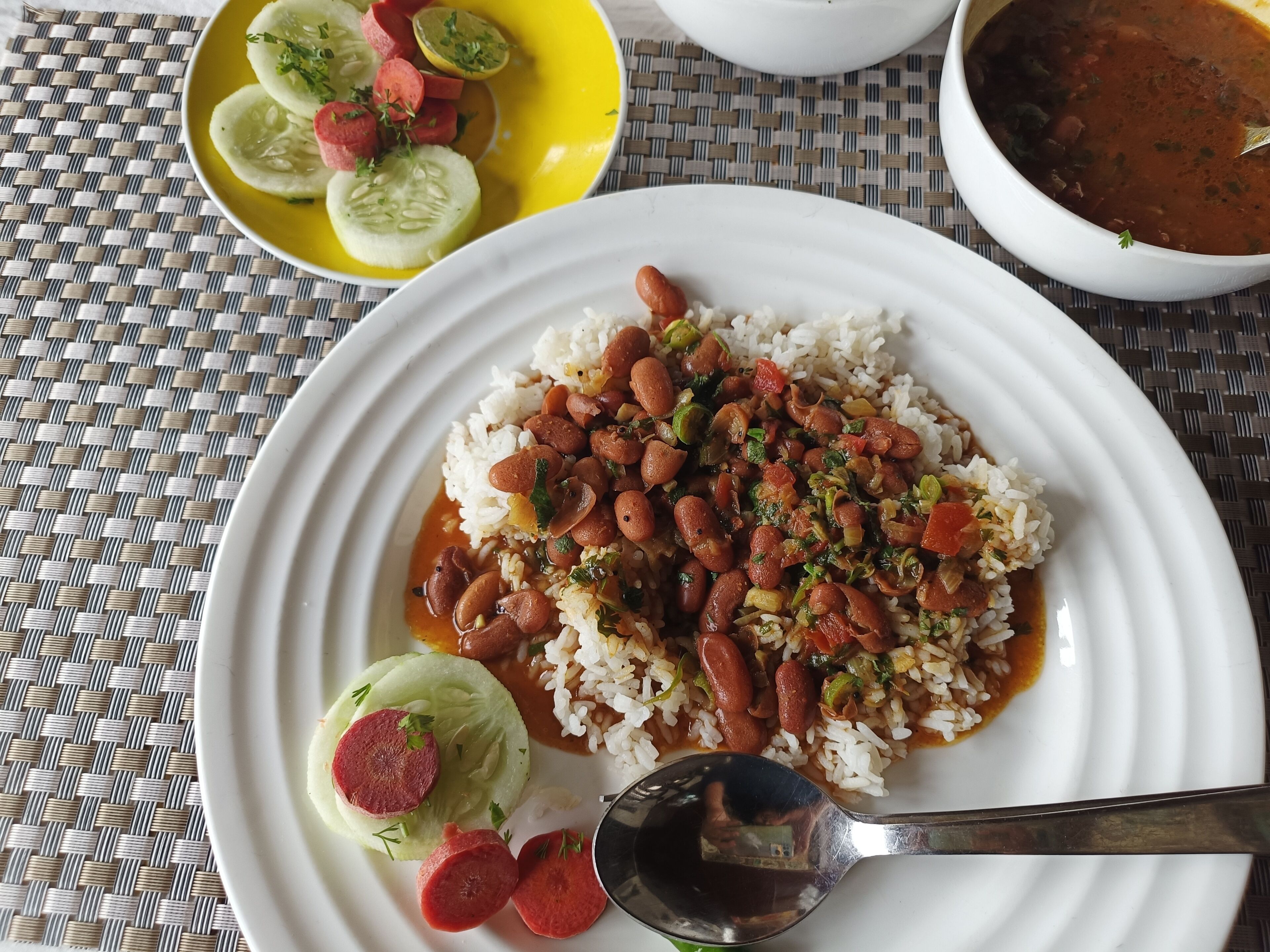 A plate of rice with beans and vegetables, a small bowl of soup, and a plate of sliced cucumbers and sausage.