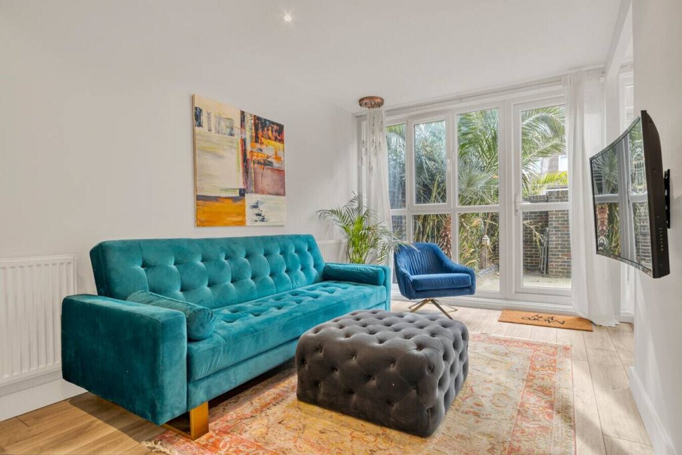 A teal sofa, a black ottoman, and a blue chair in a room with large windows and a painting on the wall.