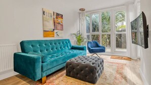A teal sofa, a black ottoman, and a blue chair in a room with large windows and a painting on the wall.