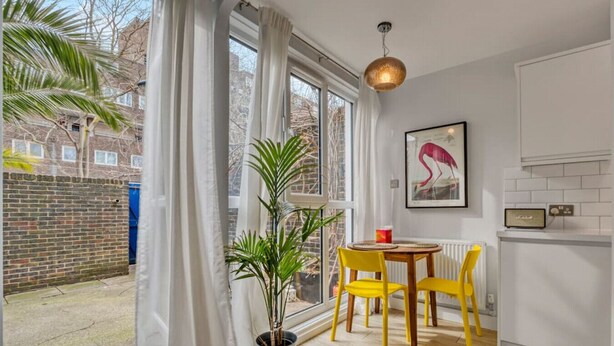 A room with a large window, a potted plant, a table with yellow chairs, and a framed artwork on the wall.