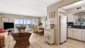 A spacious living room with a television, wicker furniture, a dining area with chairs, and a kitchen with a refrigerator.