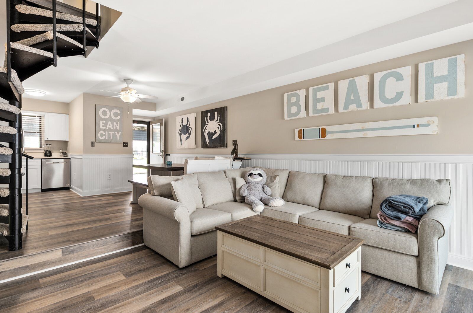 Ett rymligt vardagsrum med en beige soffa, ett träbord och en trappa med en unik design. Ett stort ord, ”BEACH”, pryder väggen, och i bakgrunden syns ett köksområde.
