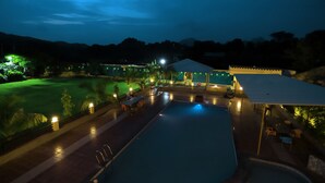 Vista nocturna de un complejo turístico con piscina, mobiliario exterior y una zona cubierta.
