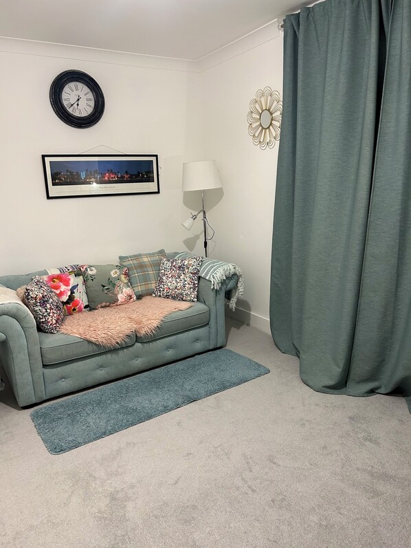 Ein Wohnzimmer mit grauem Sofa, einem gerahmten Bild, einer Uhr, einer Lampe und einem grünen Vorhang.