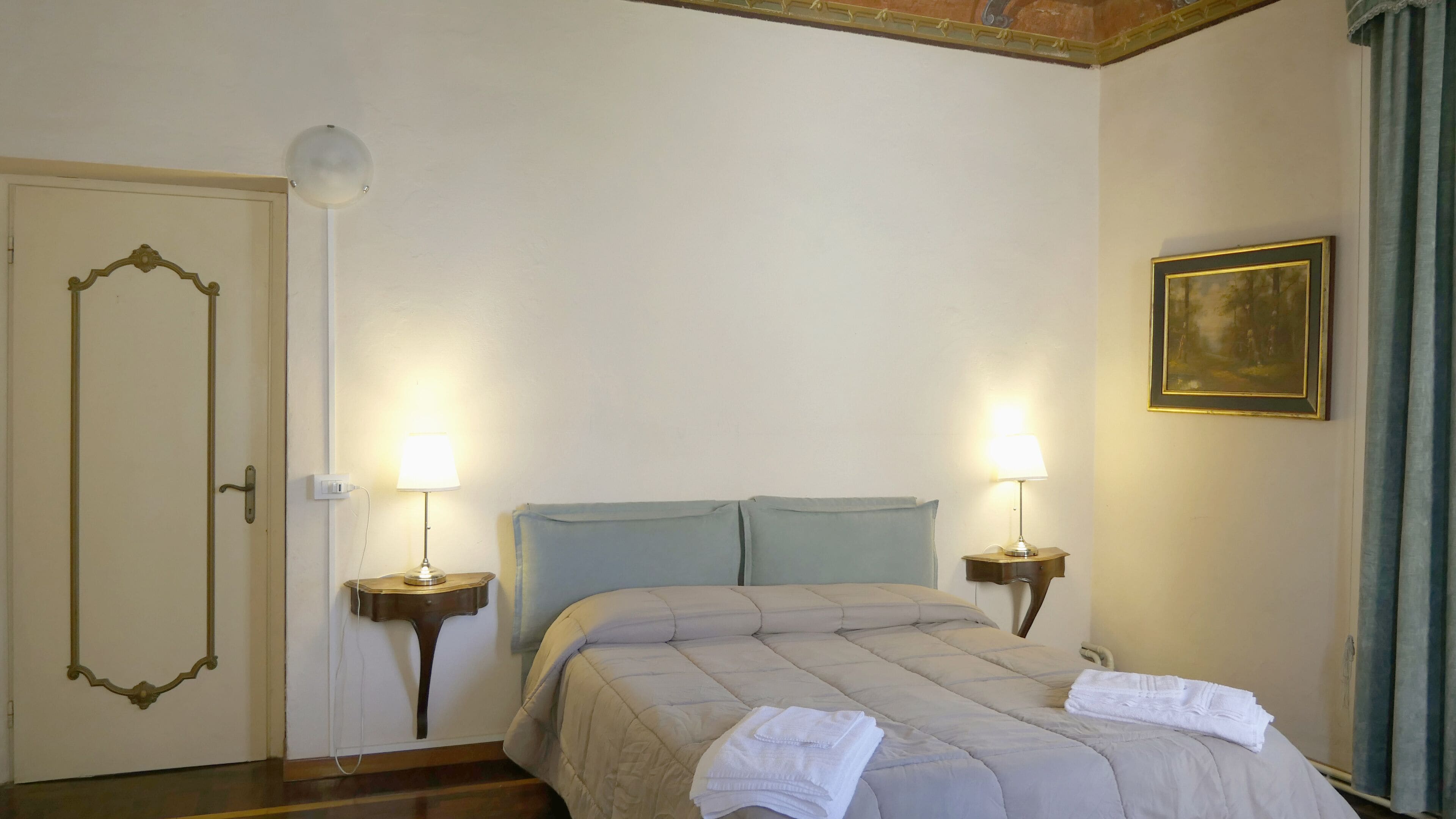 Una habitación de hotel con una cama, dos lámparas de noche, una puerta y un cuadro en la pared.