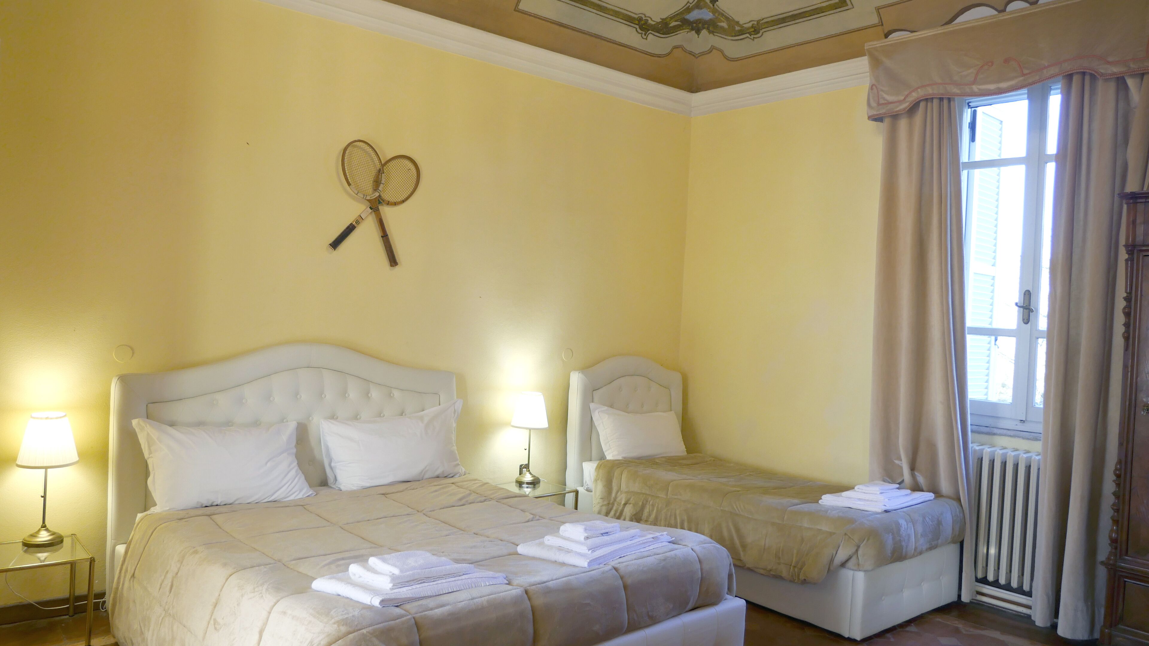 A hotel room with two beds, a window with curtains, a radiator, and a wall-mounted tennis racket.
