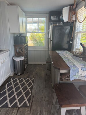 A compact kitchen with white cabinets, a wooden table, a bench, a trash can, and a window with blinds.