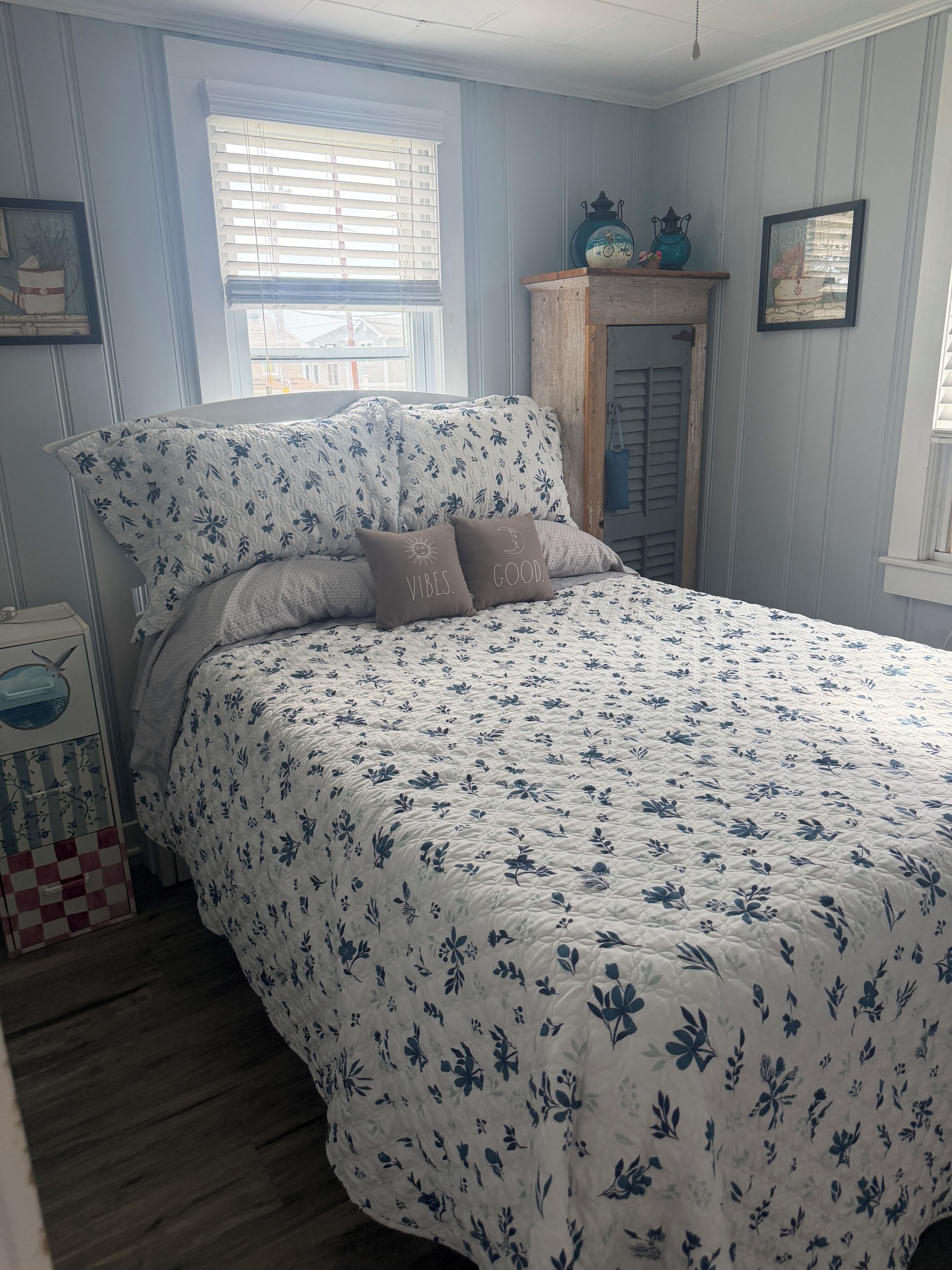 Een slaapkamer met een bloemenprint beddeken, een houten nachtkastje en een raam met zonwering.