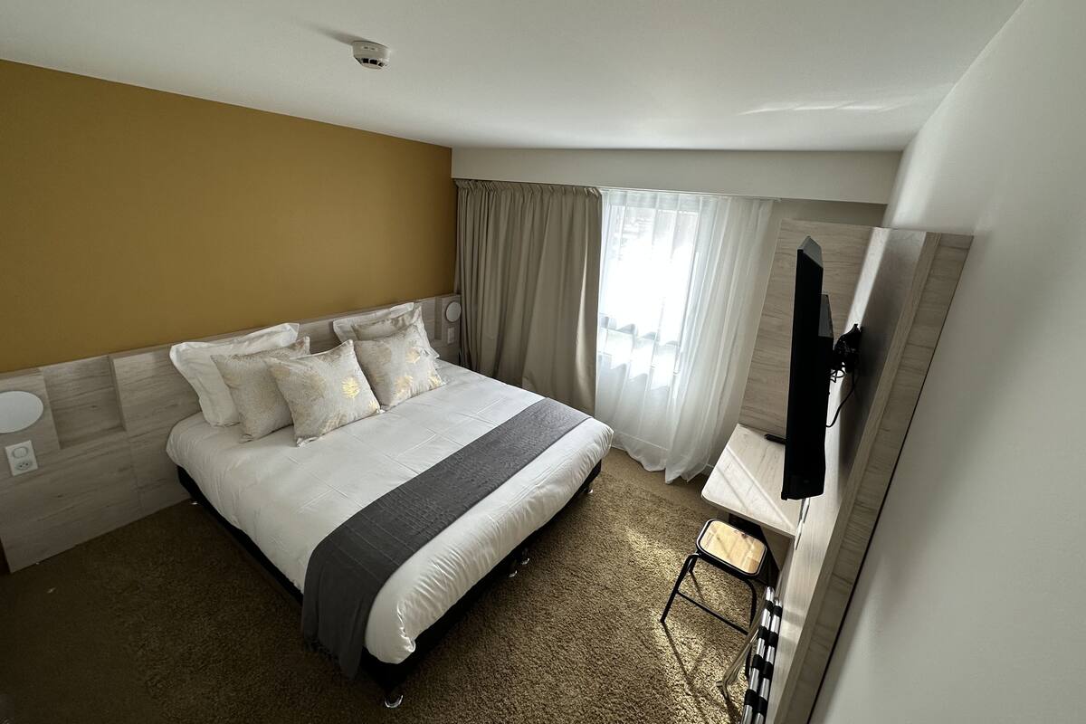 Habitación de hotel con una cama grande, una mesita de noche, un televisor montado en la pared y una ventana con cortinas.