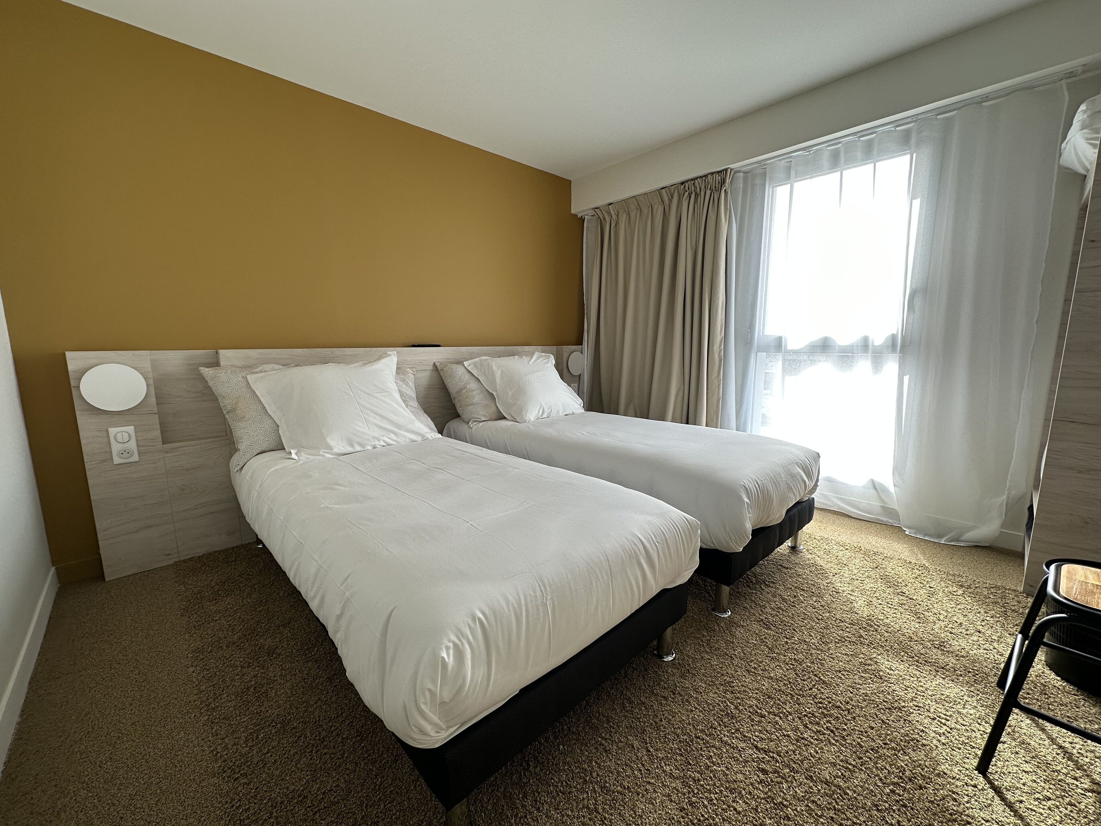 Habitación de hotel con dos camas, cabecera de madera, suelo alfombrado y ventana con cortinas.