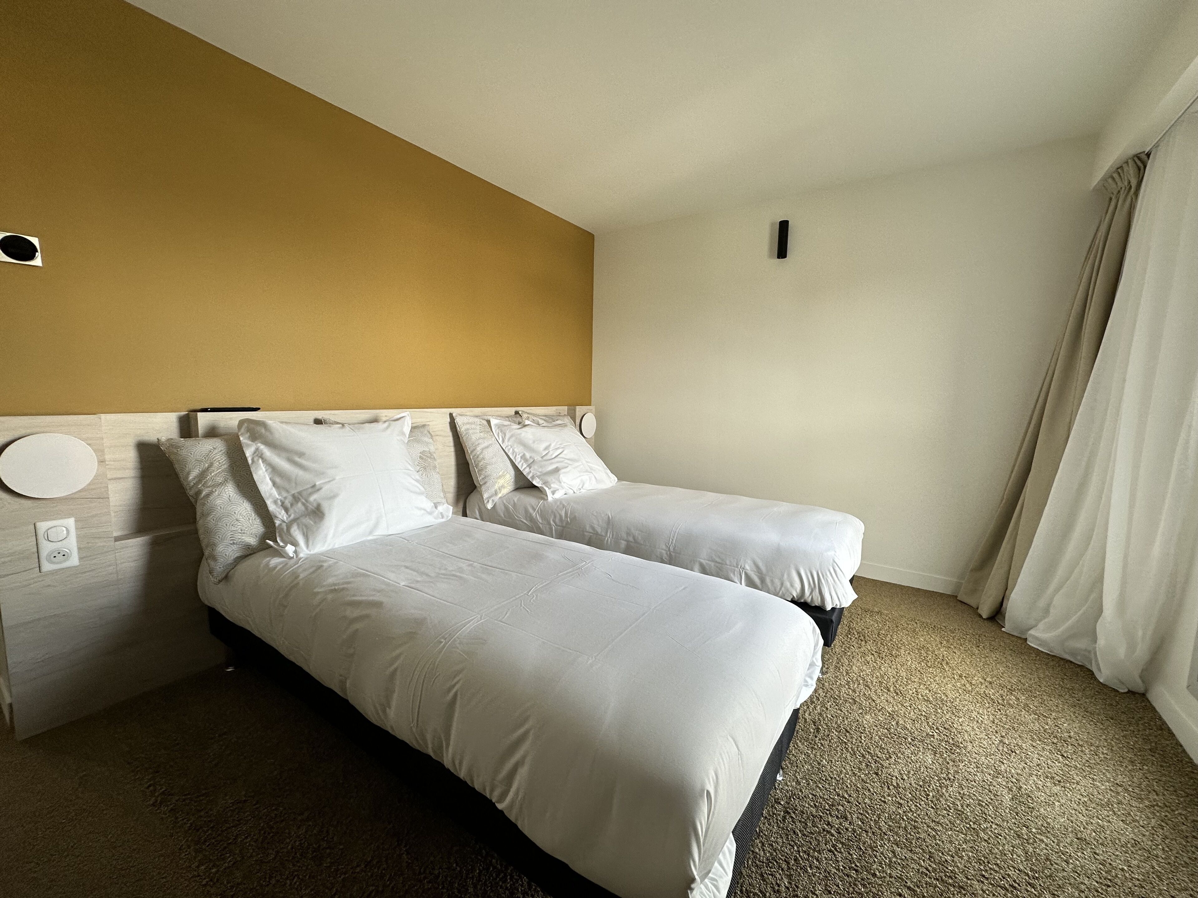 Une chambre d’hôtel avec deux lits, une tête de lit en bois, un luminaire rond et une prise de courant.