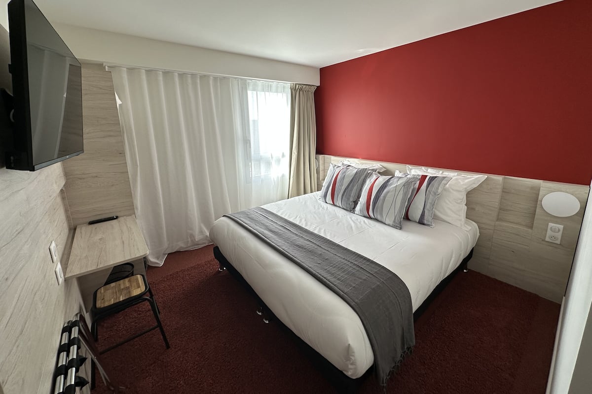 Habitación de hotel con cama, televisión montada en la pared, escritorio con silla y ventana con cortinas.