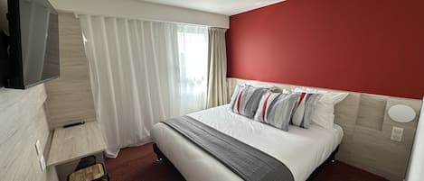 Habitación de hotel con cama, televisión montada en la pared, escritorio con silla y ventana con cortinas.