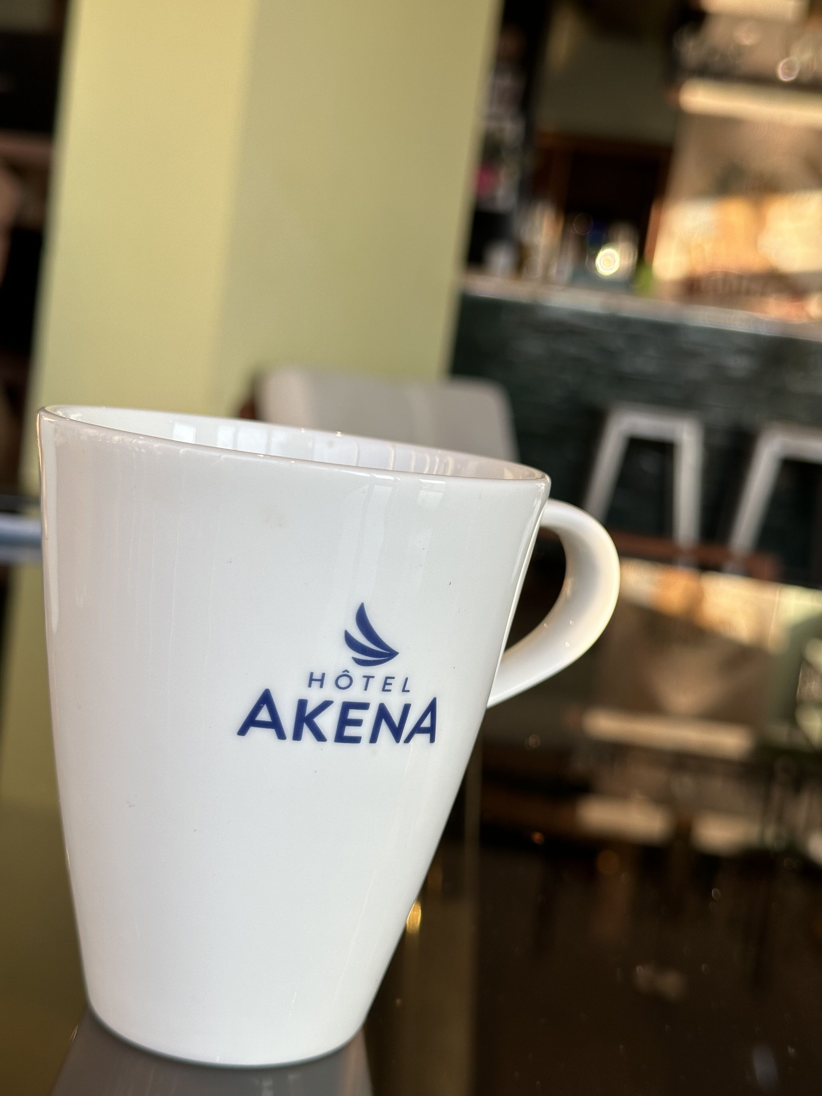 Una taza de cerámica blanca con el logotipo "HOTEL AKENA" sobre una superficie reflectante.