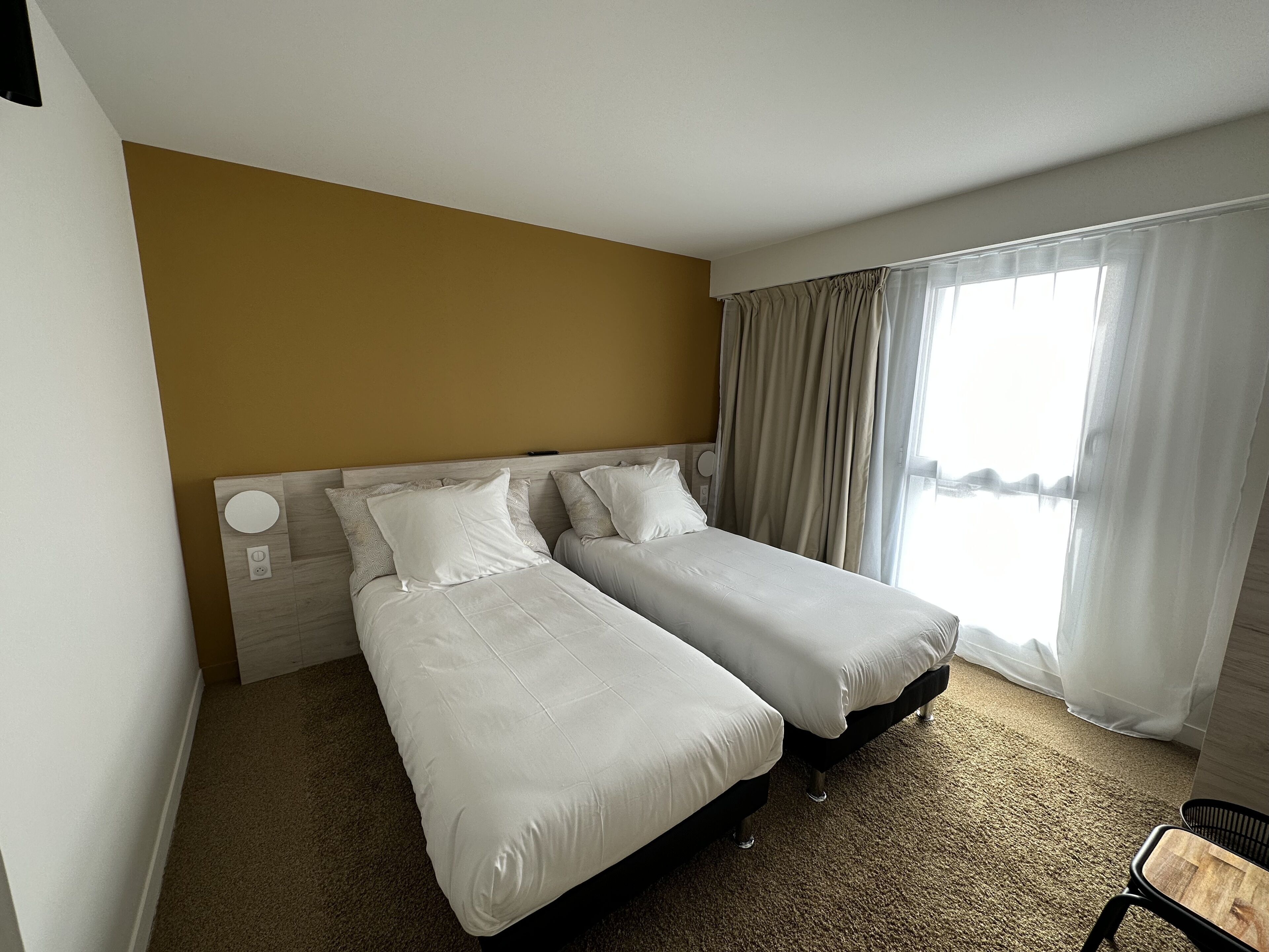 A hotel room with two single beds, a large window with curtains, a carpeted floor, and a wooden headboard.