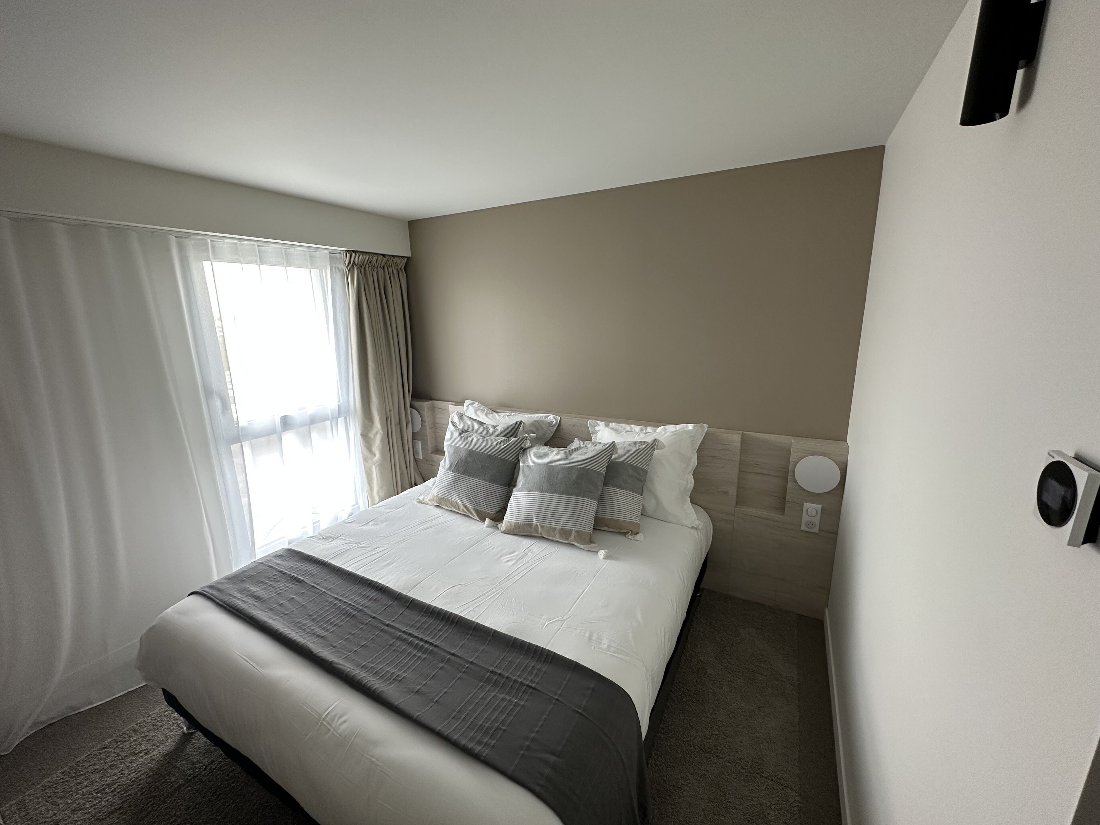 A hotel room with a large bed, a headboard, a window with curtains, and a wall-mounted air conditioning unit.