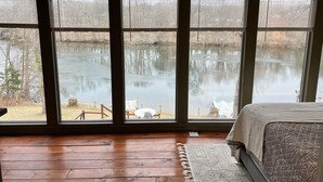 A bedroom with a large window overlooking a lake, wooden flooring, and a bed with a patterned headboard.