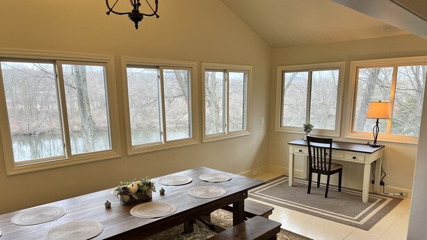 A dining area with a long wooden table, multiple windows, and a desk with a lamp.