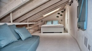 Una camera da letto con soffitto spiovente, un letto con lenzuola azzurrine, un divano bianco con cuscini blu e una TV a parete.