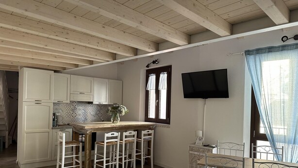 Una cucina con mobili bianchi, zoccolo di piastrelle, un tavolo da pranzo in legno con sedie, una TV montata a parete e una finestra con tende.