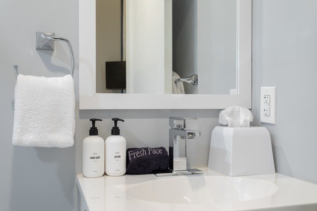 Un bagno con un lavandino bianco, uno specchio, un porta asciugamani, un dispenser per sapone liquido, una bottiglia di lozione, un asciugamano nero e una scatola di fazzoletti.