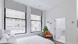 Een moderne hotelkamer met een groot bed, een fauteuil, een plant en stadszicht door grote ramen.