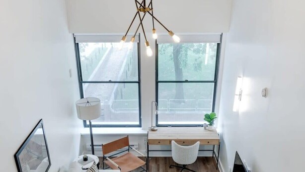 A modern living room with a sofa, coffee table, and a desk. There are large windows, a hanging light fixture, and a patterned rug.