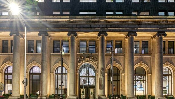 Un imposant bâtiment doté de grandes colonnes et de fenêtres en arc de cercle, sur lequel on peut lire l’inscription « JOURNAL PUBLIC ».