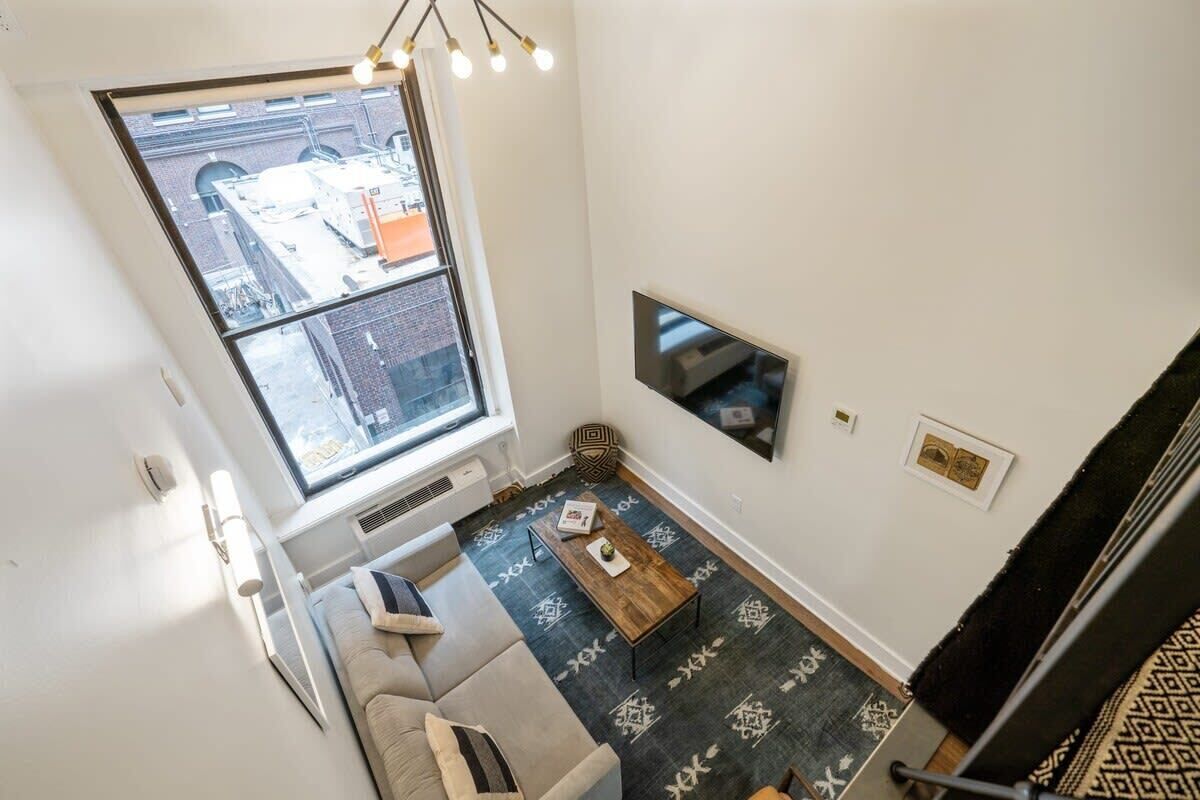 Une pièce avec un plafond haut, une grande fenêtre, un téléviseur à écran plat fixé au mur, une table basse en bois et un tapis à motifs.