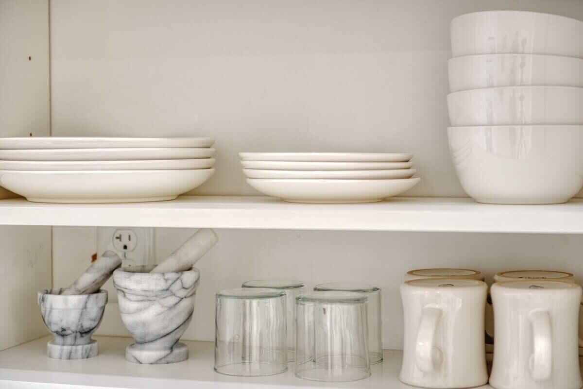 A shelf with white ceramic bowls, a marble mortar and pestle, and clear glass mugs.