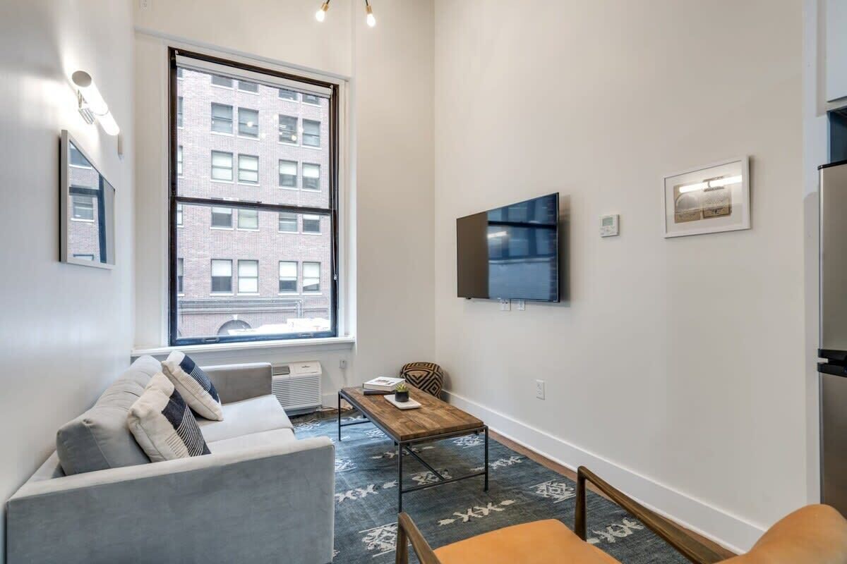 A modern living room with a grey sofa, a wooden coffee table, and a large window with a city view.